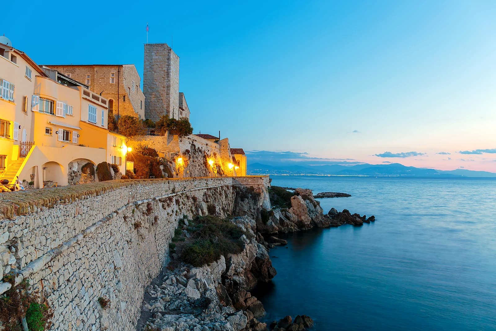 The ramparts of Old Antibes at blue hour, lamps lit above the rocks and the sea